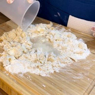 Una persona sta versando del liquido in un mucchio di farina su un tagliere.