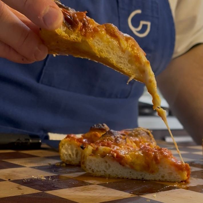 Una persona che indossa un grembiule blu con la lettera g sopra tiene in mano una fetta di pizza