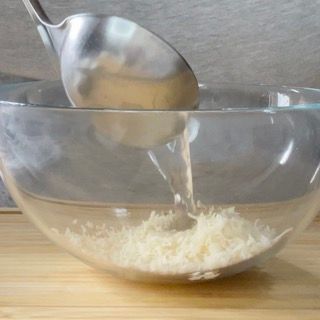Un cucchiaio versa del liquido in una ciotola di vetro piena di formaggio grattugiato.