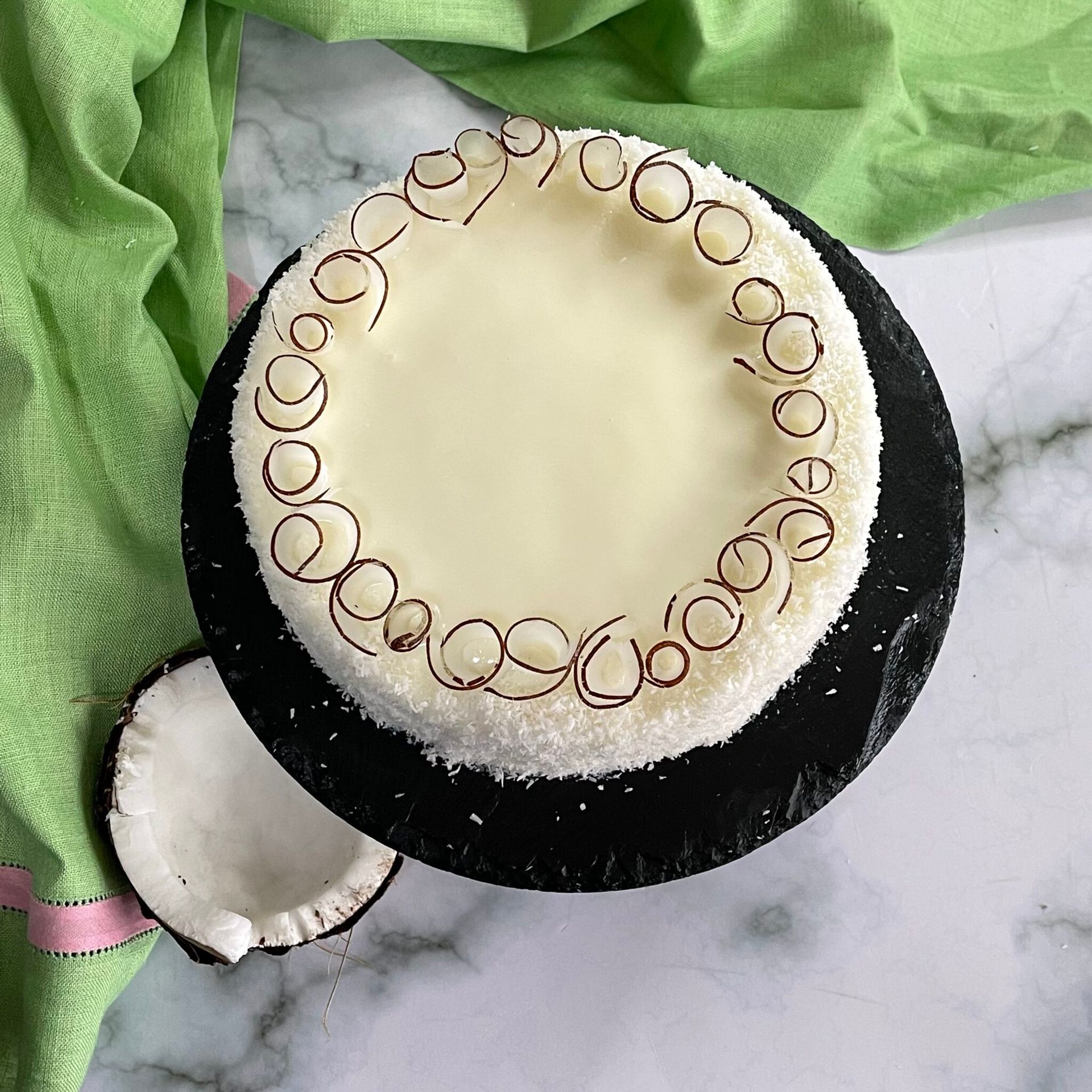 Una torta al cocco è appoggiata su un piatto nero su un tavolo accanto a una noce di cocco.