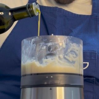 Versamento a filo dell'olio extravergine d'oliva sopra la purea d'aglio per iniziare l'emulsione.