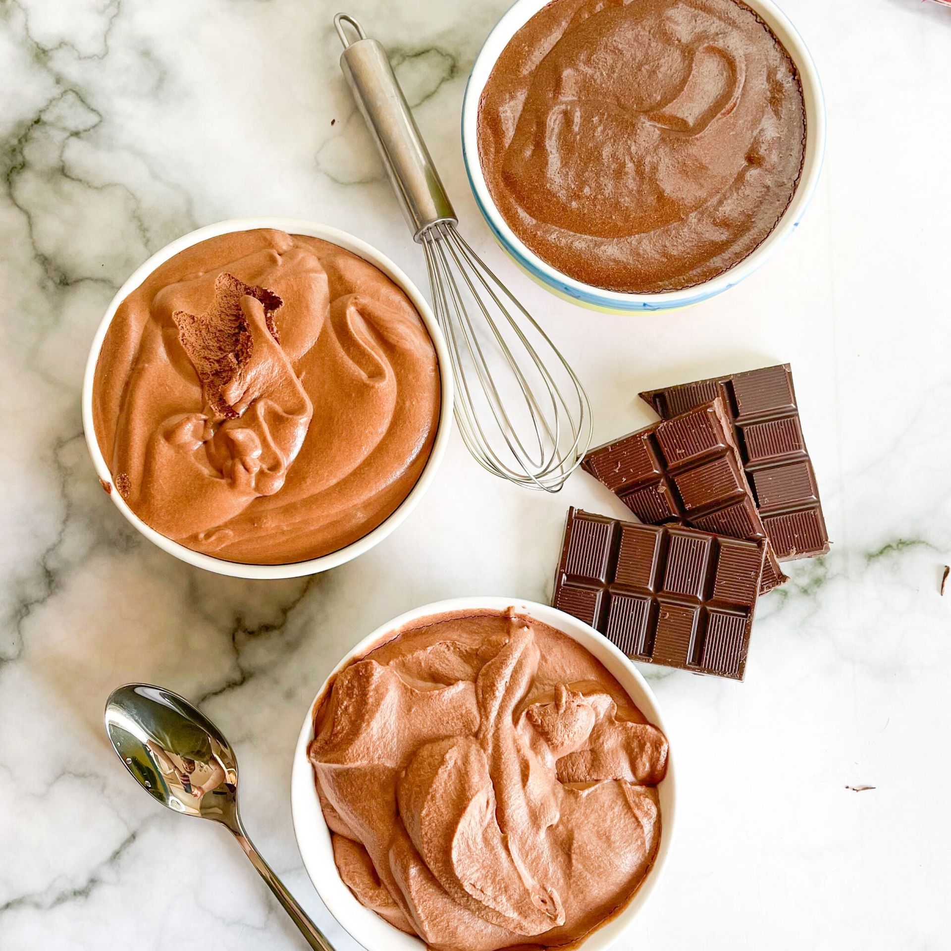 Tre ciotole di mousse al cioccolato accanto a una frusta e delle barrette di cioccolato.