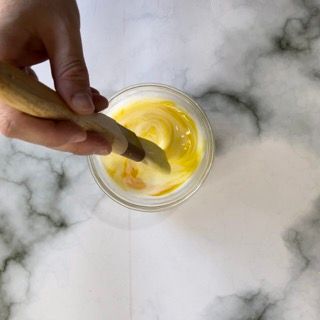 Una persona tiene in mano un cucchiaio di legno in una ciotola contenente un liquido giallo.