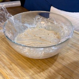 Una ciotola di vetro piena di spaghetti avvolti nella pellicola trasparente su un tavolo di legno.