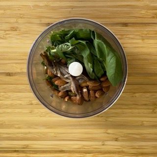 Un robot da cucina pieno di noci, basilico e altre verdure su un tavolo di legno.