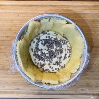 Una torta con gocce di cioccolato sopra è avvolta nella pellicola trasparente e posta su un tagliere di legno.