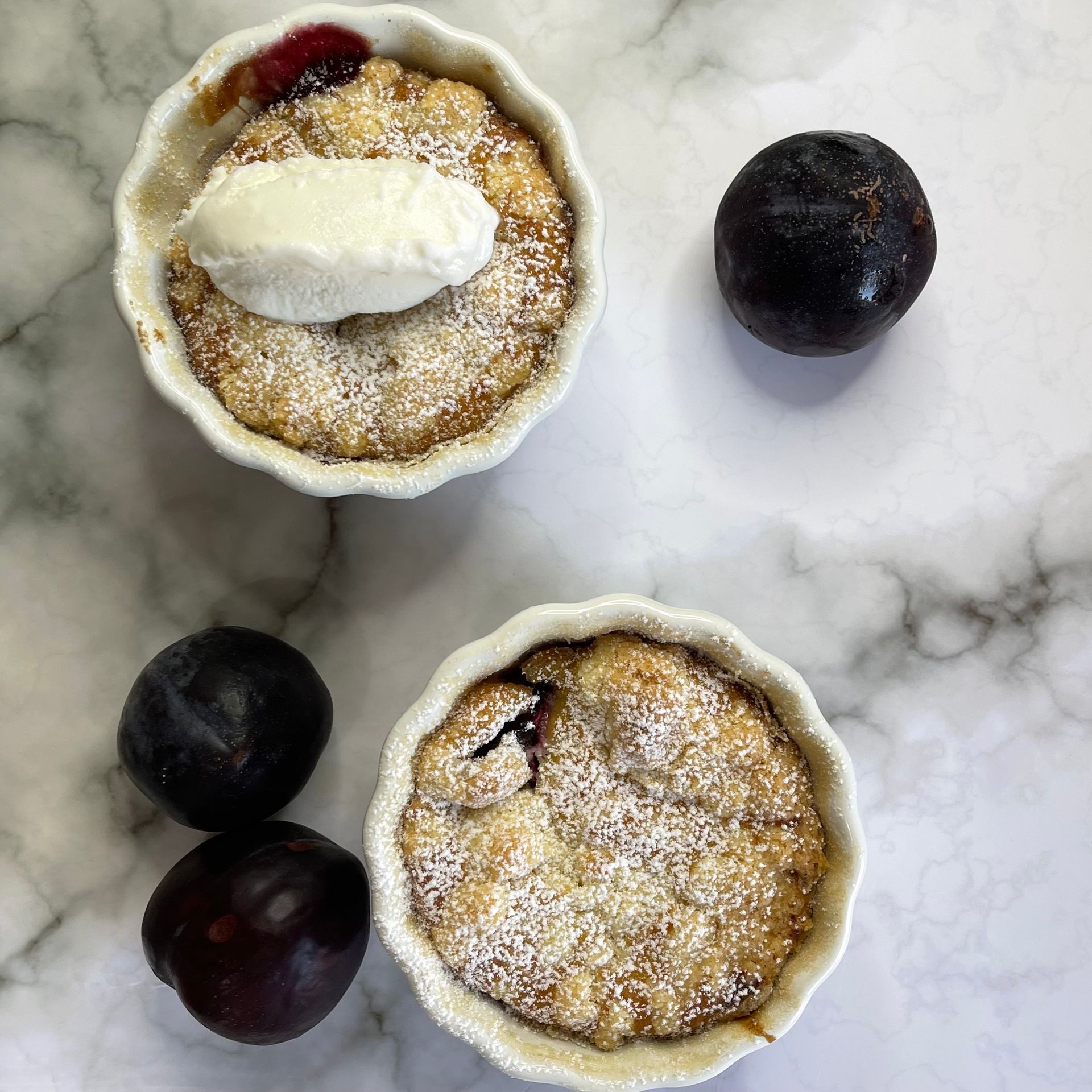Due ciotole di crumble di prugne con panna montata e prugne su un tavolo.
