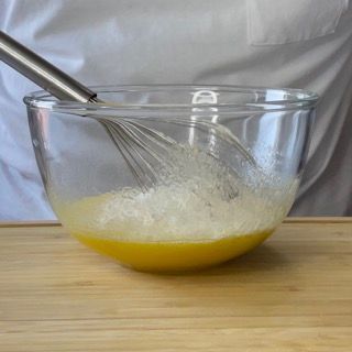 Una ciotola di vetro piena di liquido giallo e una frusta su un tavolo di legno.