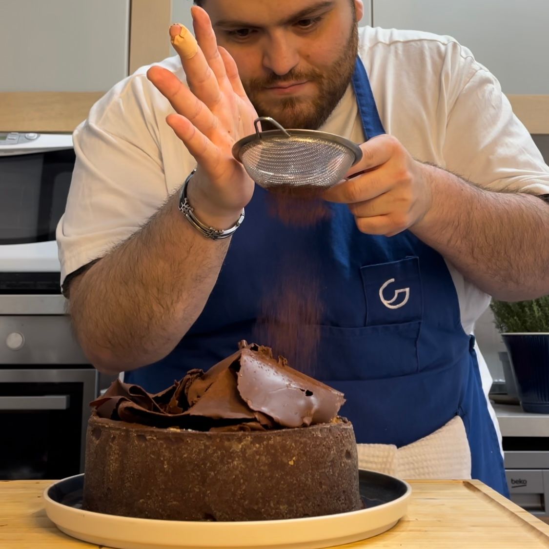 Un uomo con un grembiule blu sta spolverando una torta al cioccolato con zucchero a velo