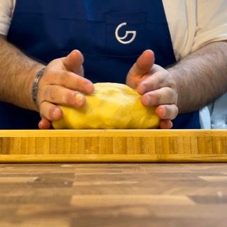 Un uomo che indossa un grembiule con la lettera g sopra tiene in mano una palla di pasta gialla