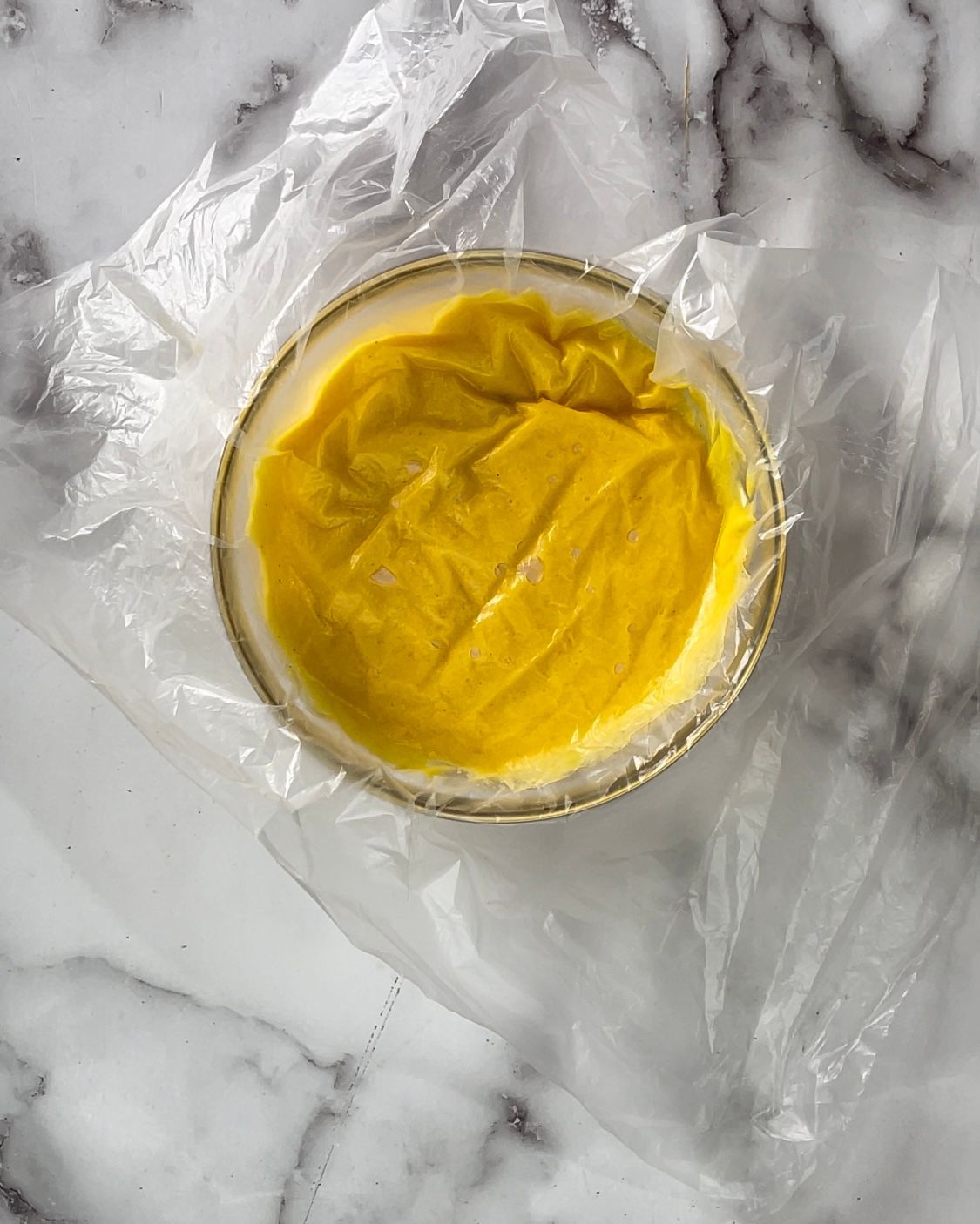 Una ciotola piena di liquido giallo avvolta nella plastica su una superficie di marmo.