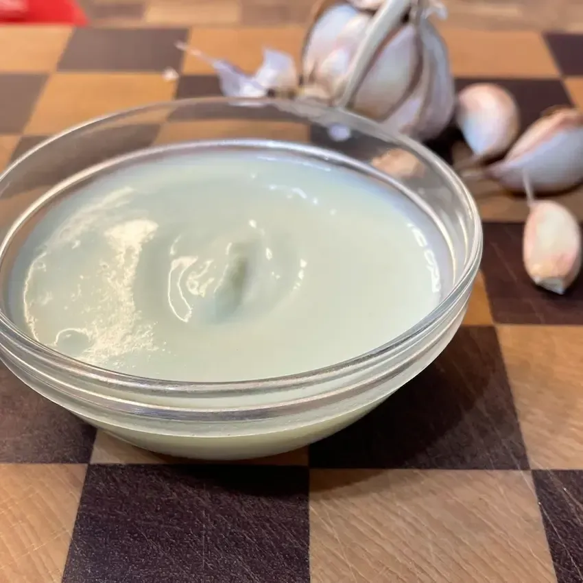 Una ciotolina di ceramica piena di crema di aglio fatta in casa, bianca, vellutata e soffice, decorata con foglioline di timo fresco e circondata da teste d'aglio intere per la crema di aglio ricetta originale.