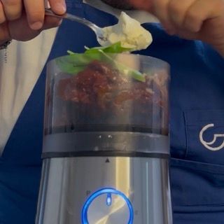 Aggiunta di foglie di basilico fresco e formaggio grattugiato nel frullatore per il pesto di pomodori secchi.