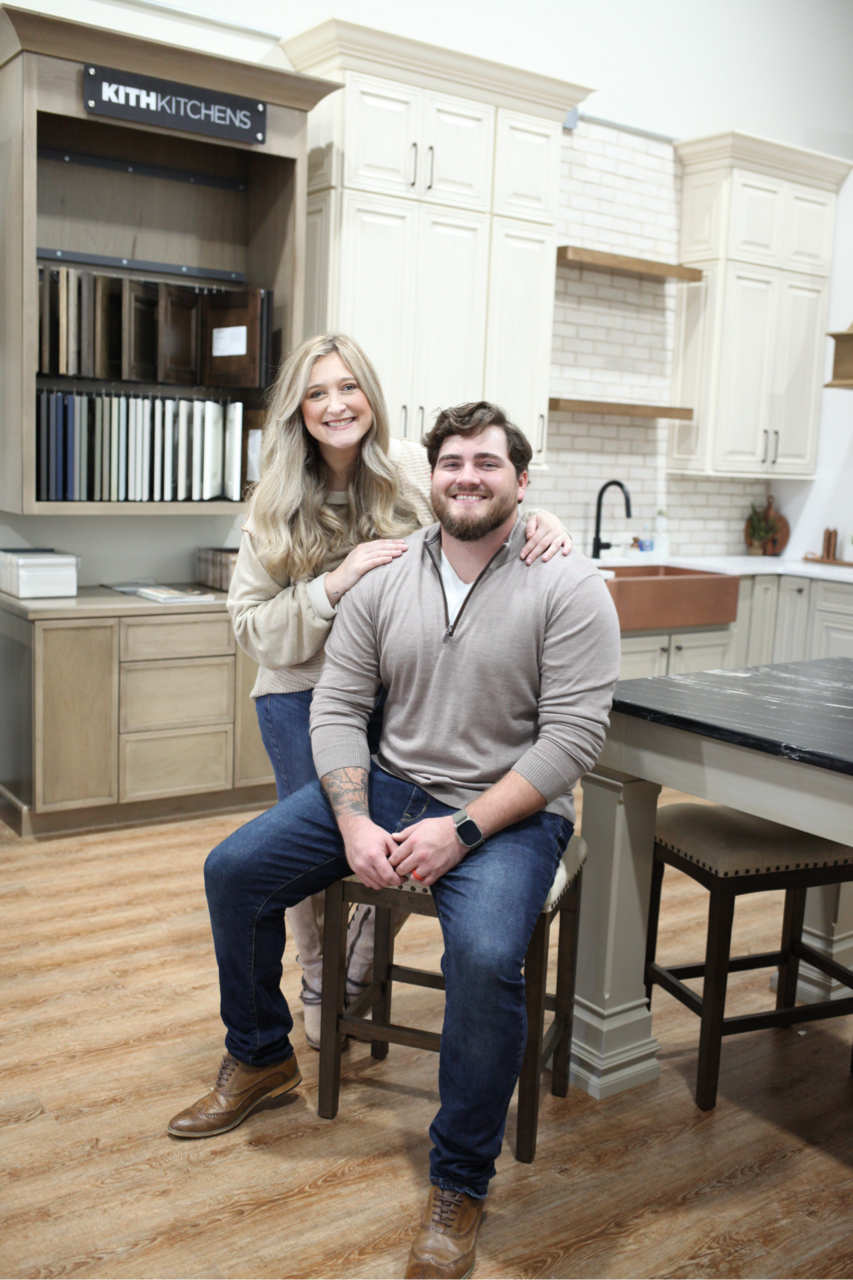 Two people posing in a bright kitchen showroom with cabinets, island, and stools.