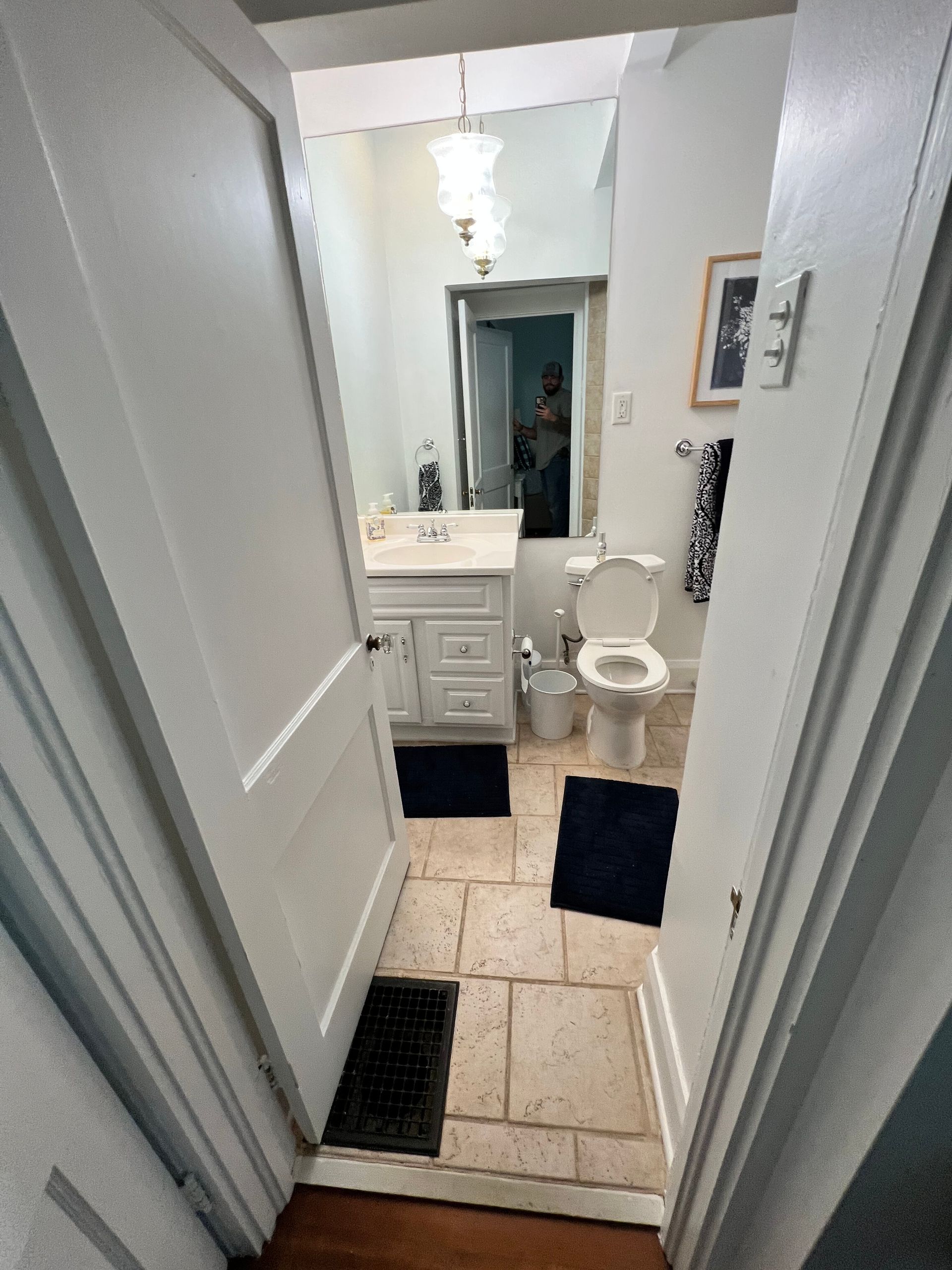 Narrow bathroom with white vanity, toilet, mirror, and dark floor mats viewed from the doorway