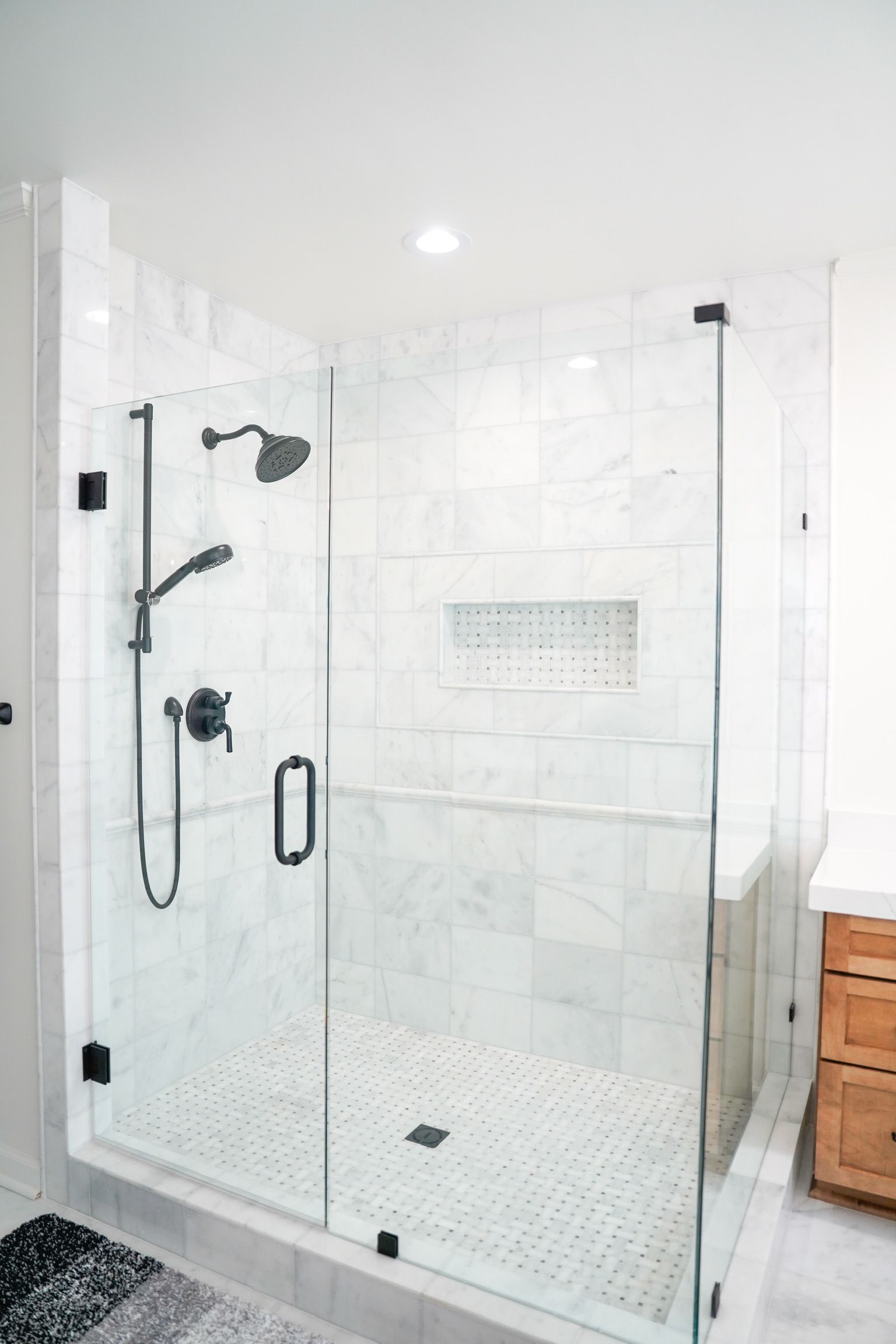 Glass walk-in shower with white tile, black fixtures, and a built-in shelf in a bright bathroom