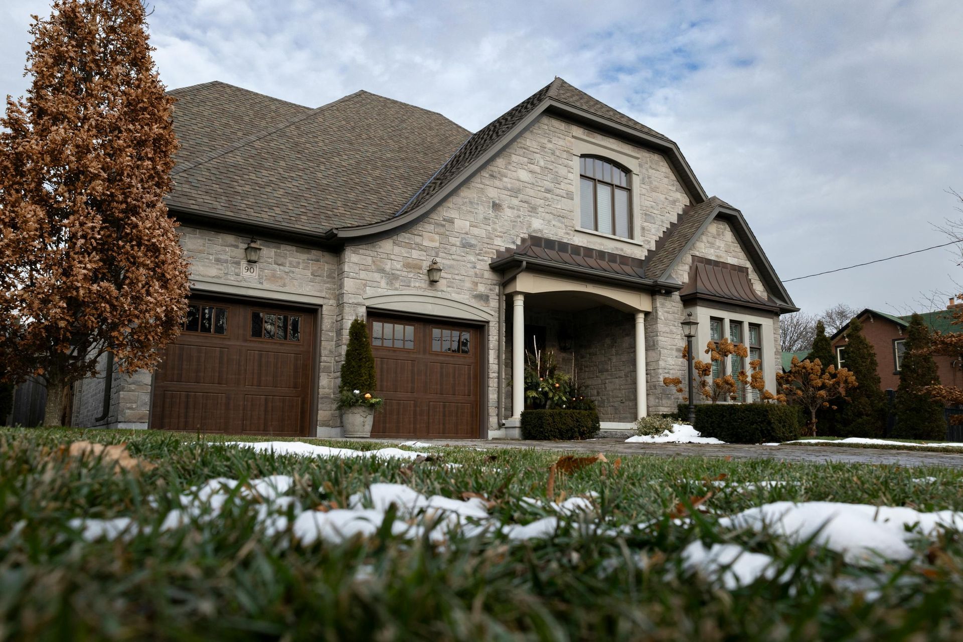 Stone suburban house with garage and snow-dusted lawn under a cloudy sky