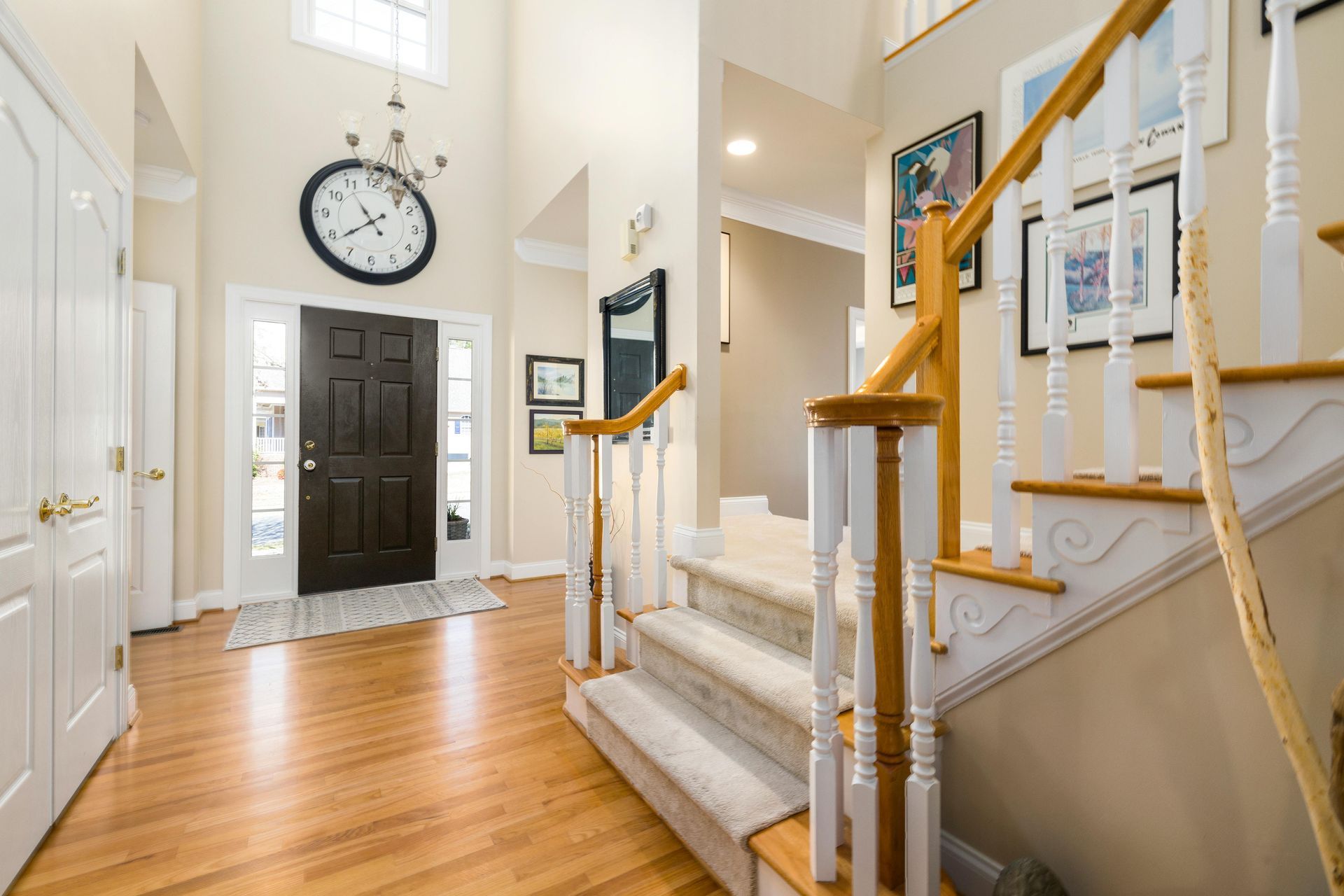 Bright foyer with hardwood floors, white staircase, black front door, and large wall clock