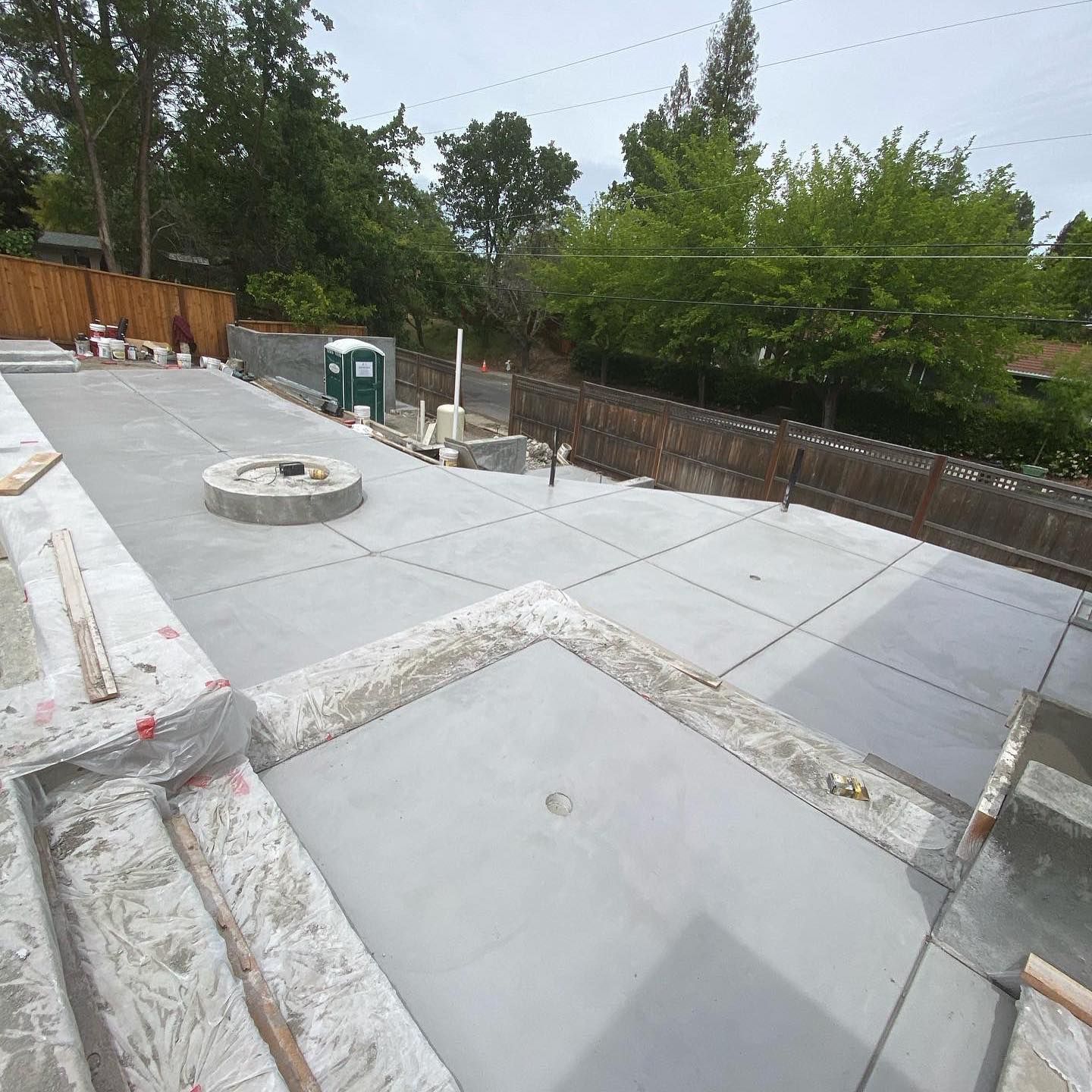 Freshly poured concrete patio with a circular fire pit base, surrounded by wooden fencing in a backyard setting.