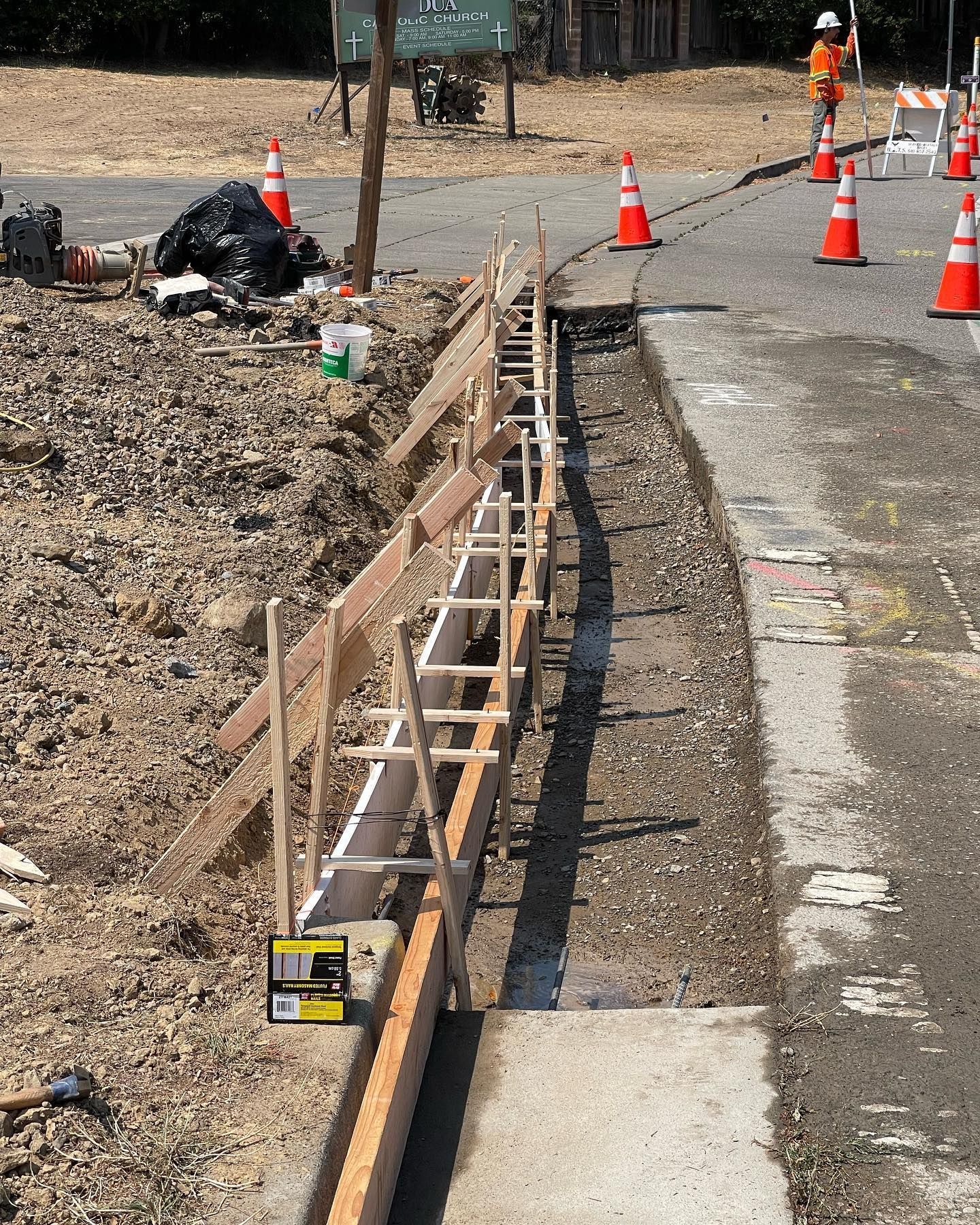 Wooden construction forms for a new curb line along an asphalt road with traffic cones and a worker in the background.