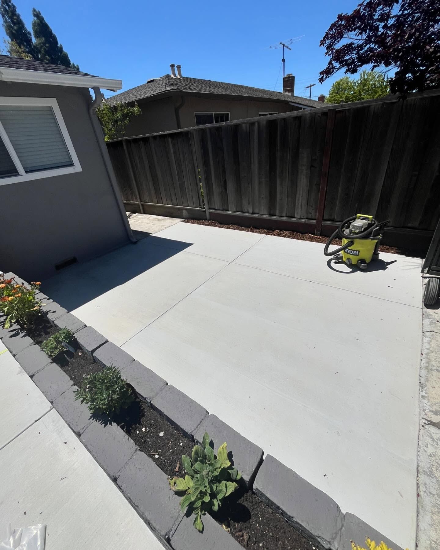 A newly poured concrete patio in a residential backyard, bordered by a dark stone planter and a wooden fence.