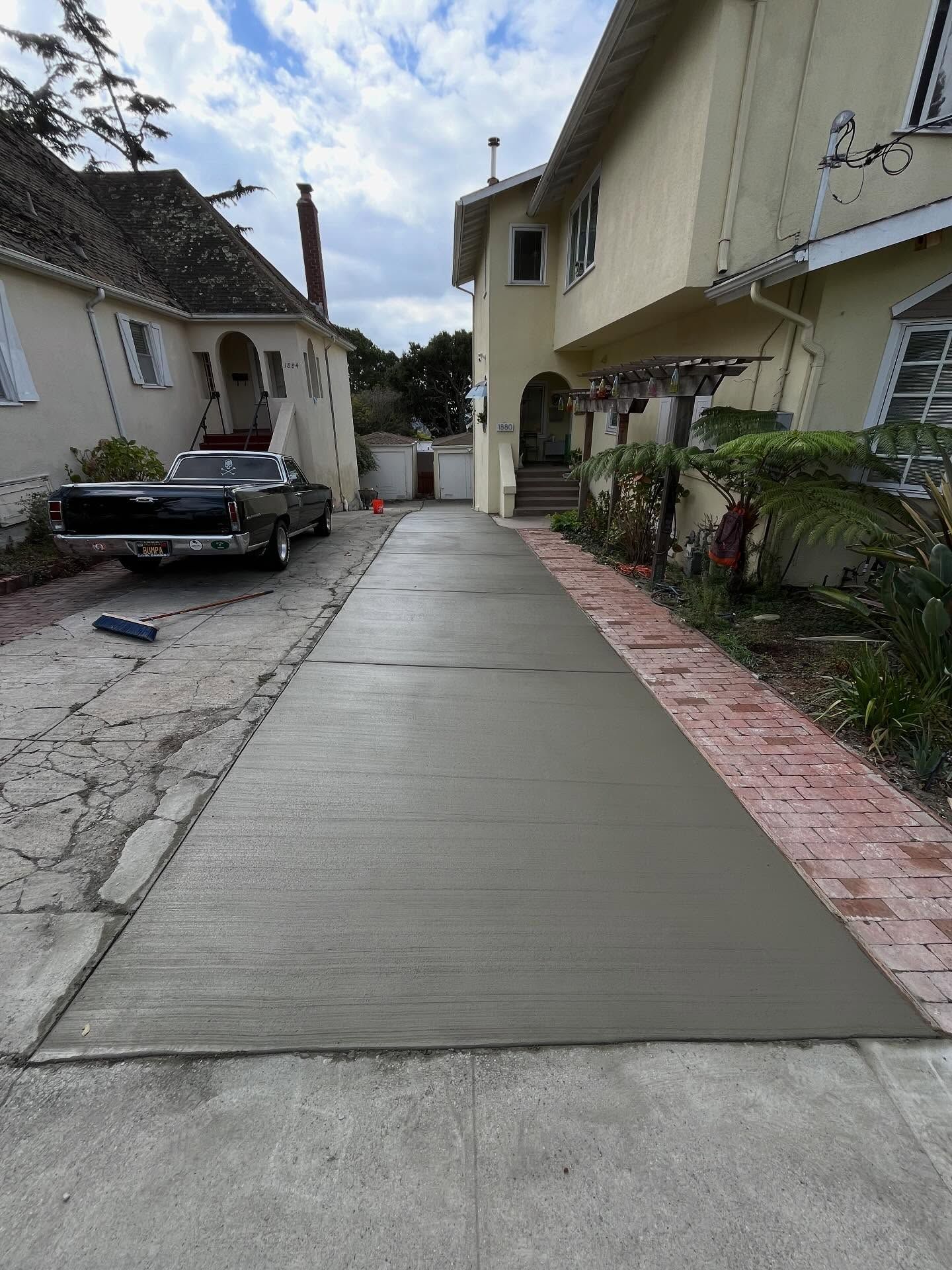 A freshly poured concrete driveway section sits between two houses, with a black truck parked on the left.