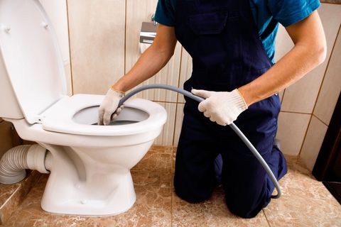 Plumber using a drain snake to unclog a toilet in a bathroom, wearing gloves and blue uniform