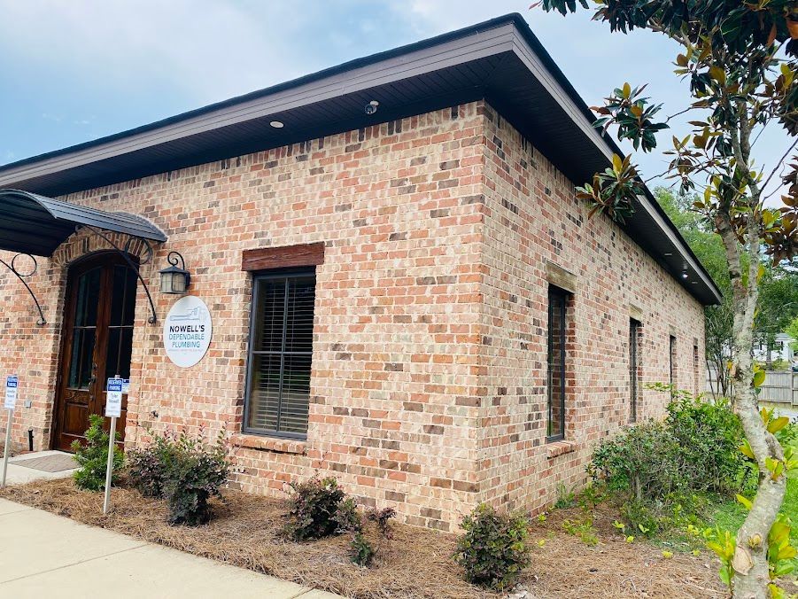 Brick building with a dark roof, barred windows, and a small entrance sign amid shrubs and a sidewalk