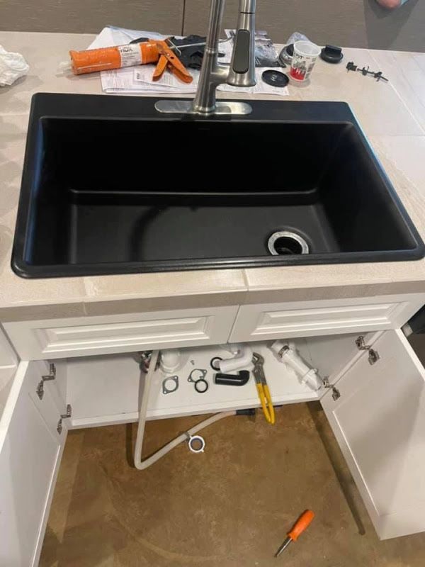 Bathroom vanity with black sink and open cabinet showing plumbing tools and supplies inside.