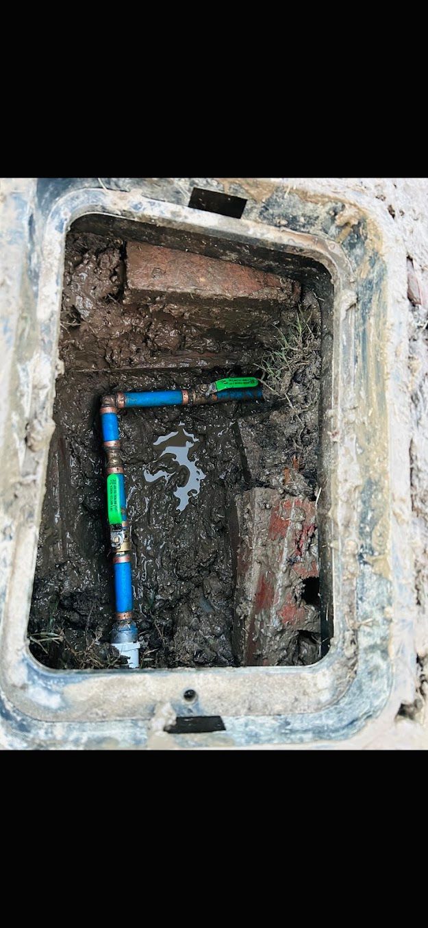 Open utility box with exposed multicolored pipes and muddy, debris-filled interior