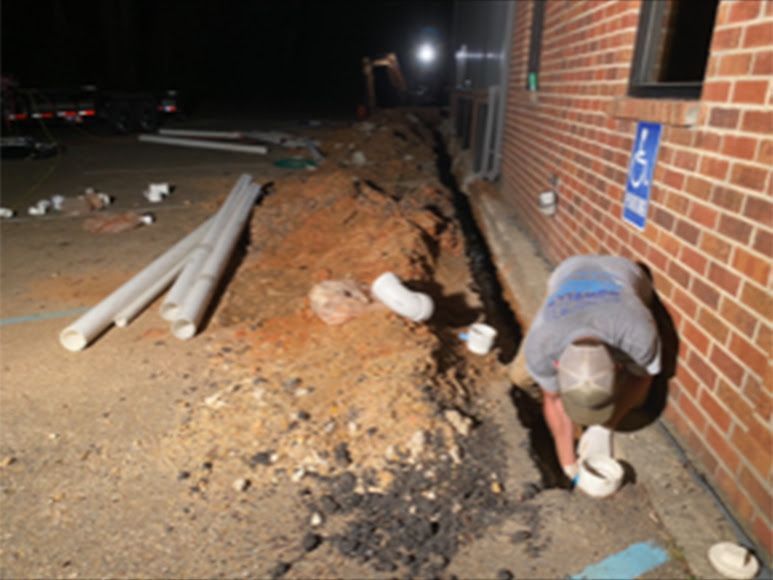 Worker digging a trench beside a brick building at night, with pipes and dirt piles along the wall.