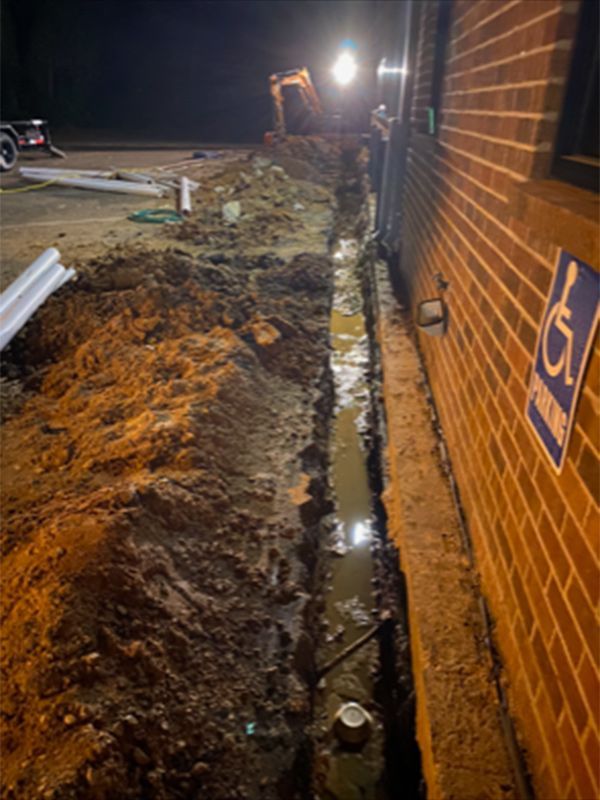 Nighttime construction trench beside brick wall, with excavator and muddy dirt pile under work lights