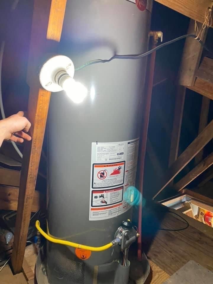 Water heater in an attic with a lit bulb, yellow gas line, and warning labels.