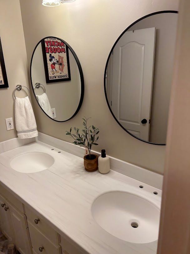 Bright bathroom vanity with double sinks, round mirrors, beige walls, and a white door reflected in one mirror