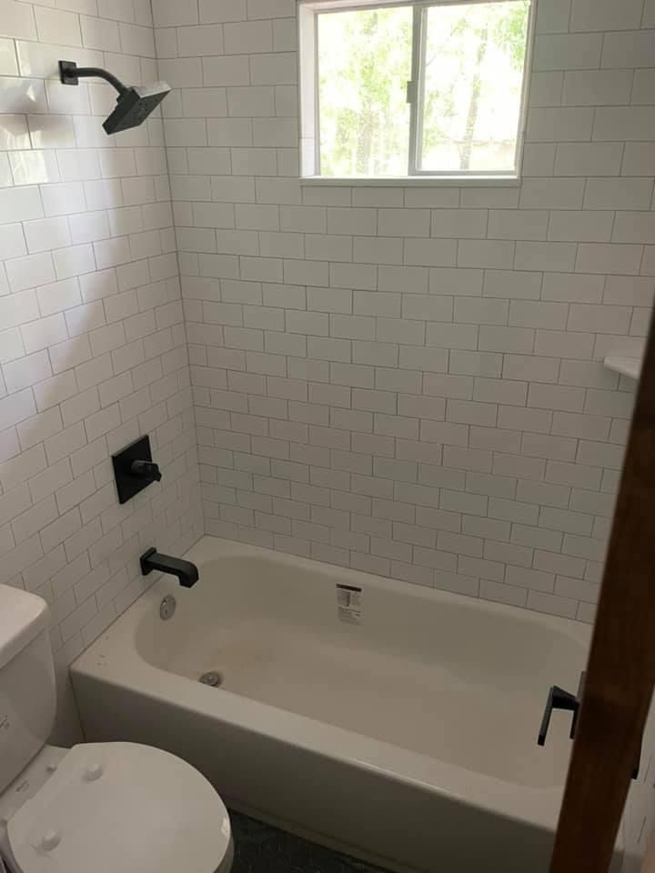 Small bathroom with white subway-tile walls, tub, toilet, window, and black fixtures.
