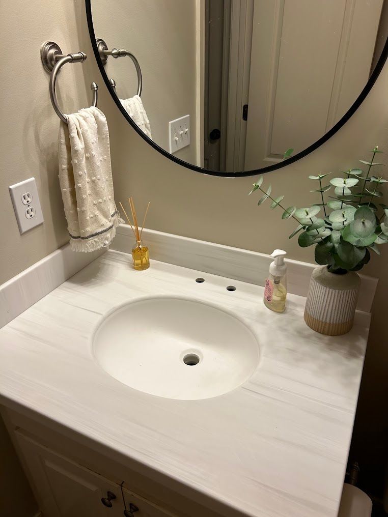 Bathroom vanity with round mirror, white sink, towel, soap dispenser, and vase of greenery.