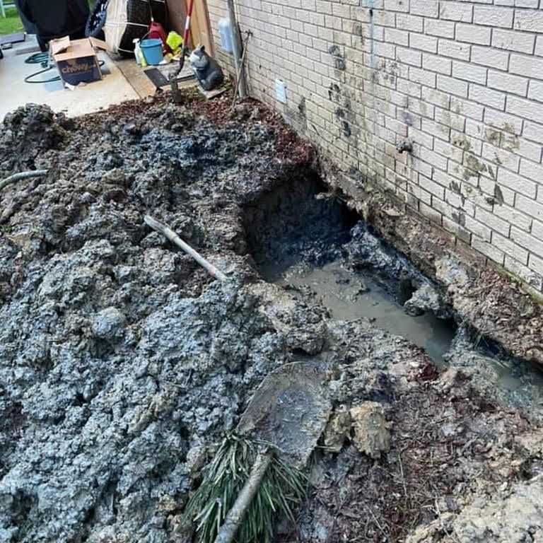 Flood-damaged yard beside a brick wall, with muddy debris and a large hole exposed near the foundation.