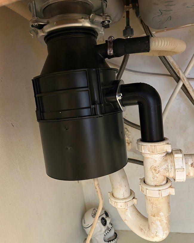 Black garbage disposal under a sink with white drain pipes and a hose connection.