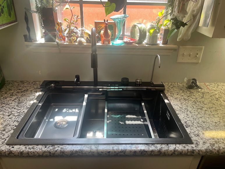 Kitchen sink with black basin, granite countertop, and window with plants above it.