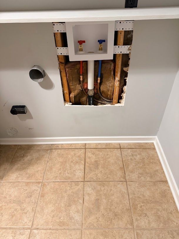 Laundry room wall with open plumbing access panel above beige tile floor