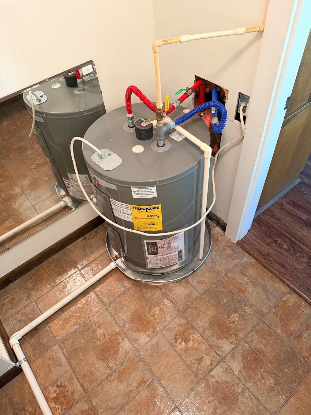 Water heater with red and blue pipes beside a wall and mirror on a tiled floor