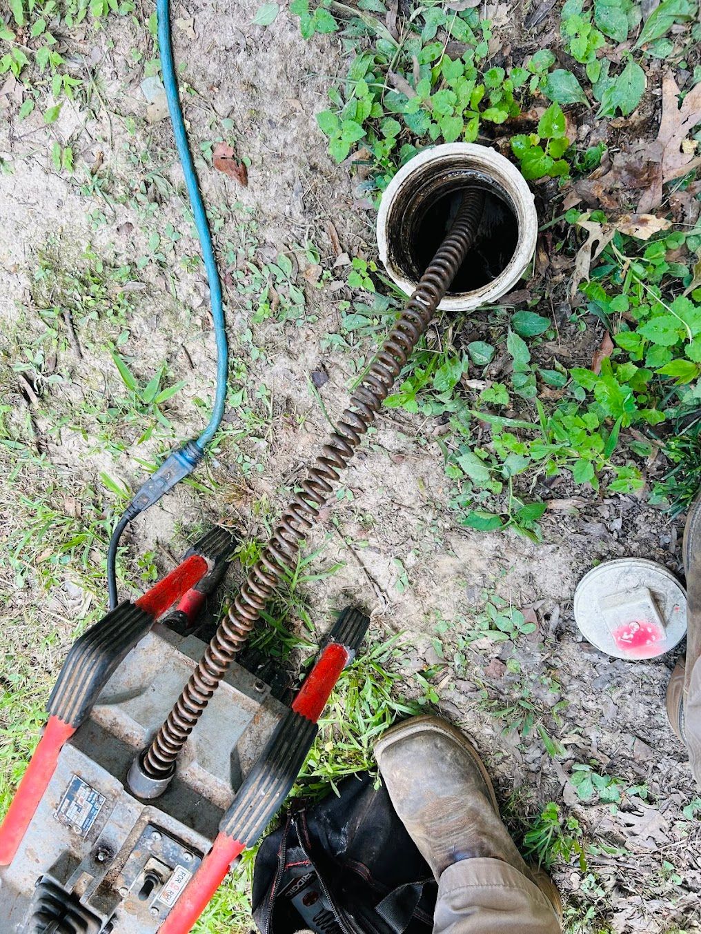 Drain snake entering outdoor pipe beside a hose and boot on dirt and weeds