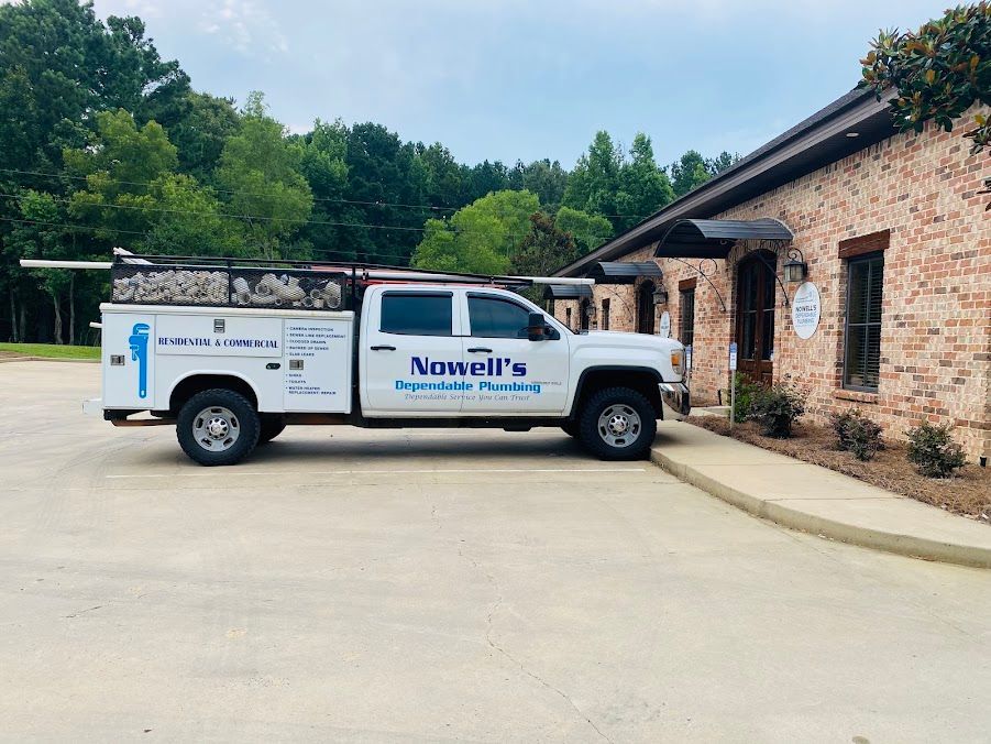 White Novelis truck parked by a stone building on a concrete lot with trees in the background