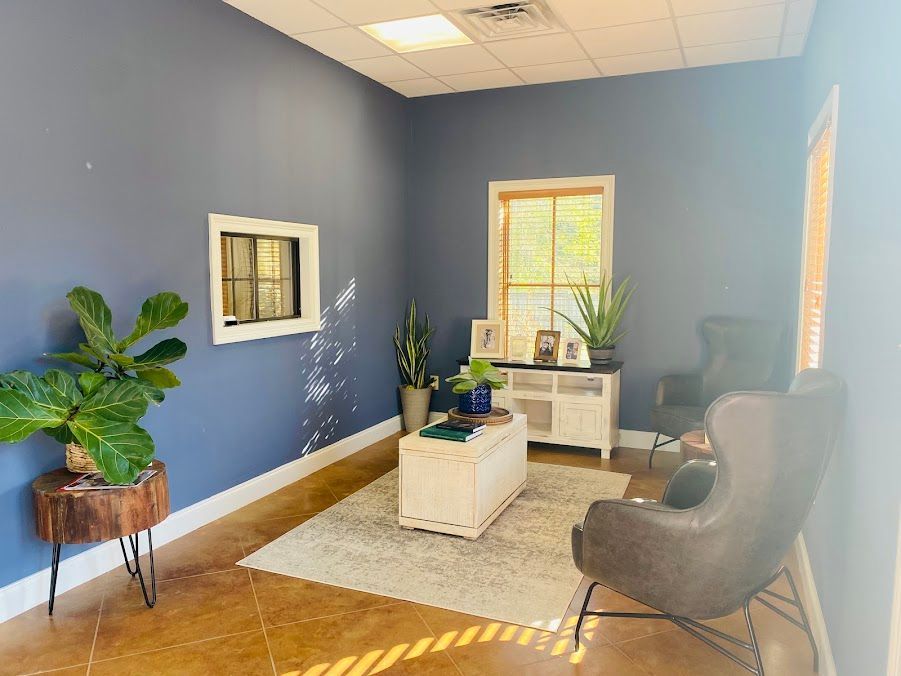 Blue waiting room with gray chairs, white tables, plants, and sunlight from windows.