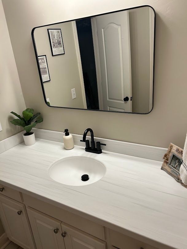 Modern bathroom vanity with white countertop, black faucet, round sink, mirror, plant, and decor.