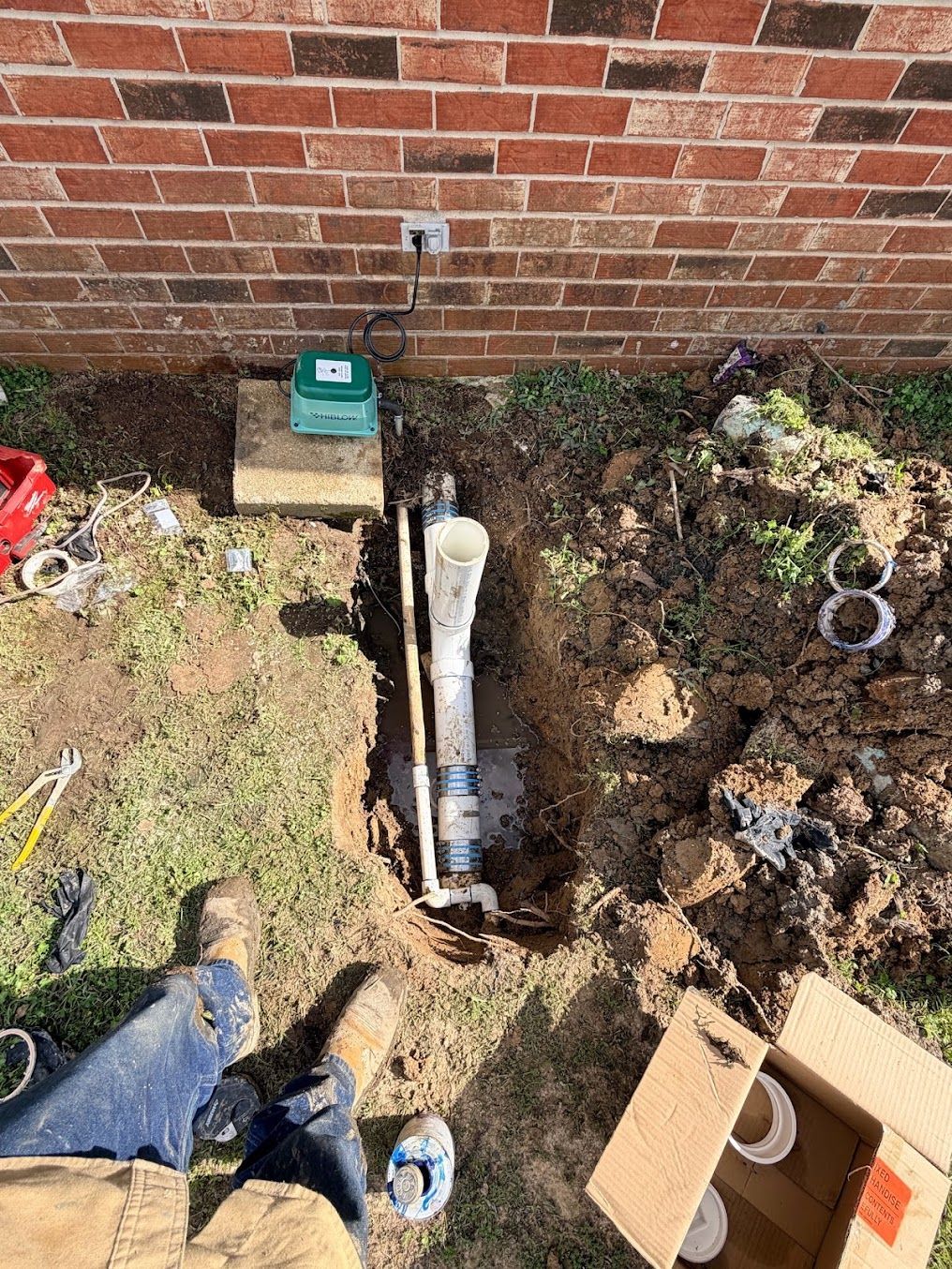 Open trench beside brick wall with exposed pipe and utility box in a yard