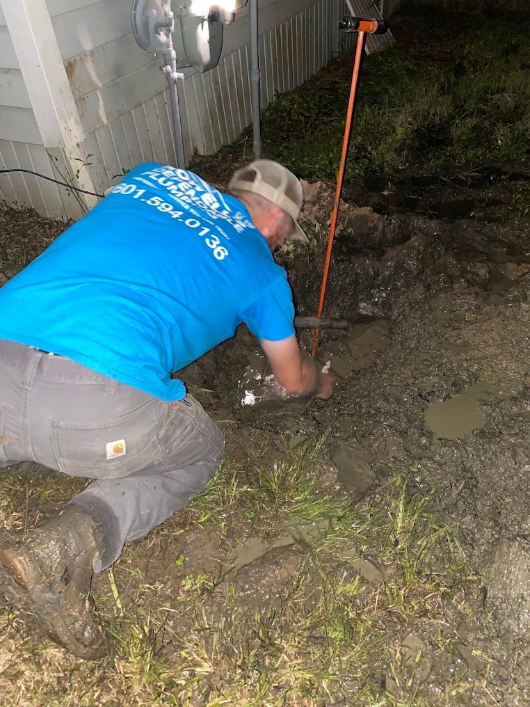 Worker kneeling by a crawlspace opening, inspecting muddy ground beneath a house with tools nearby.