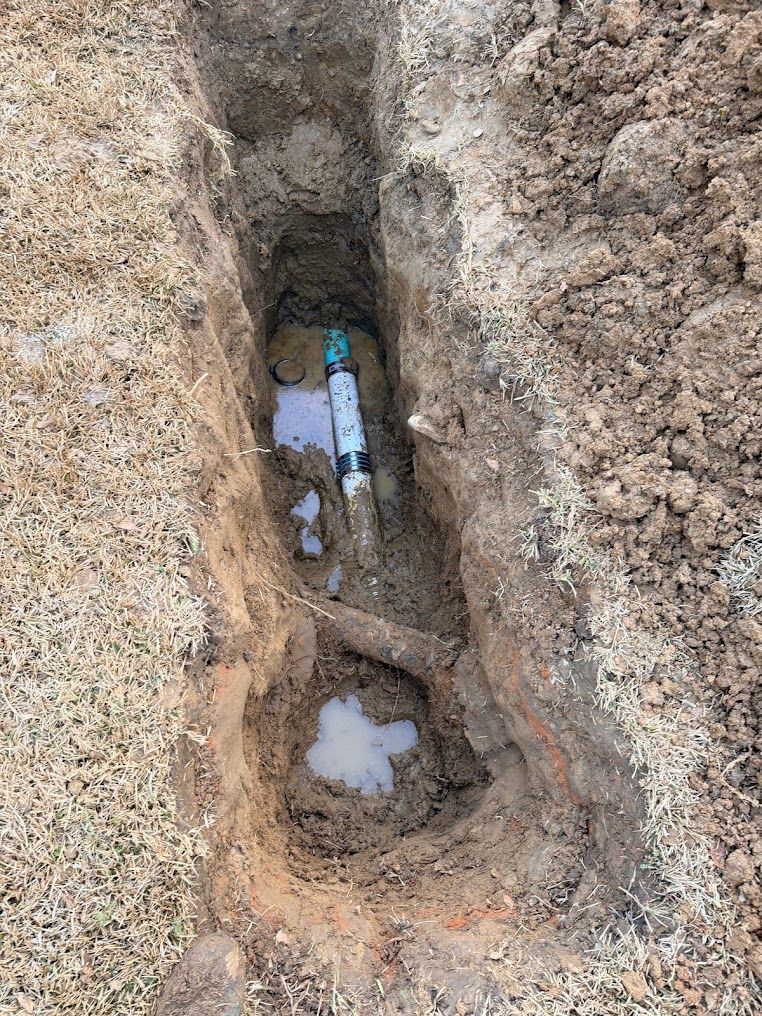 Excavated trench exposing underground pipes in soil, with muddy water pooled at the bottom