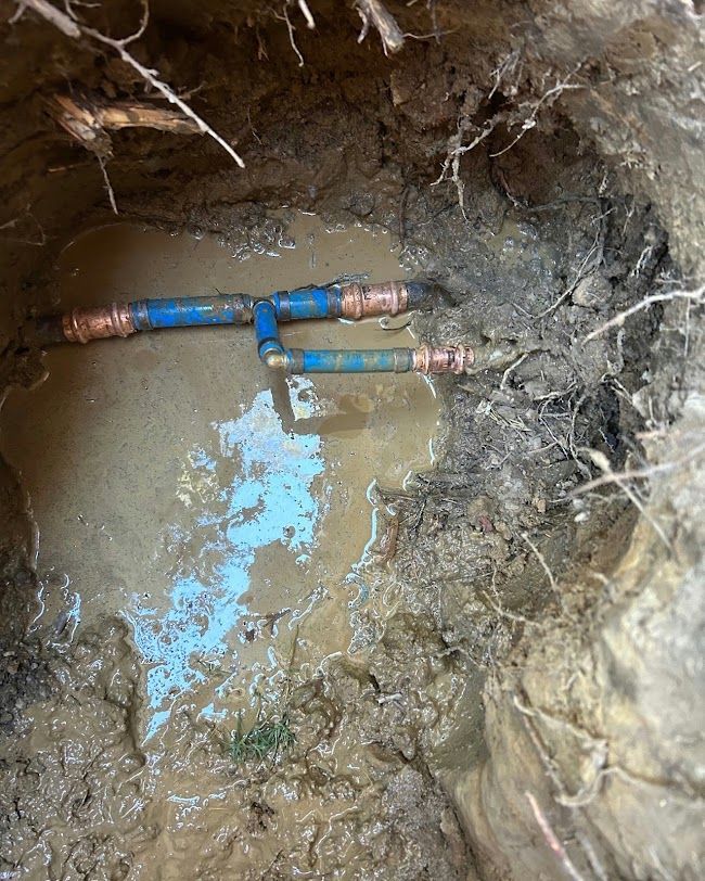 Exposed underground blue pipes in a muddy trench with standing water and dirt walls