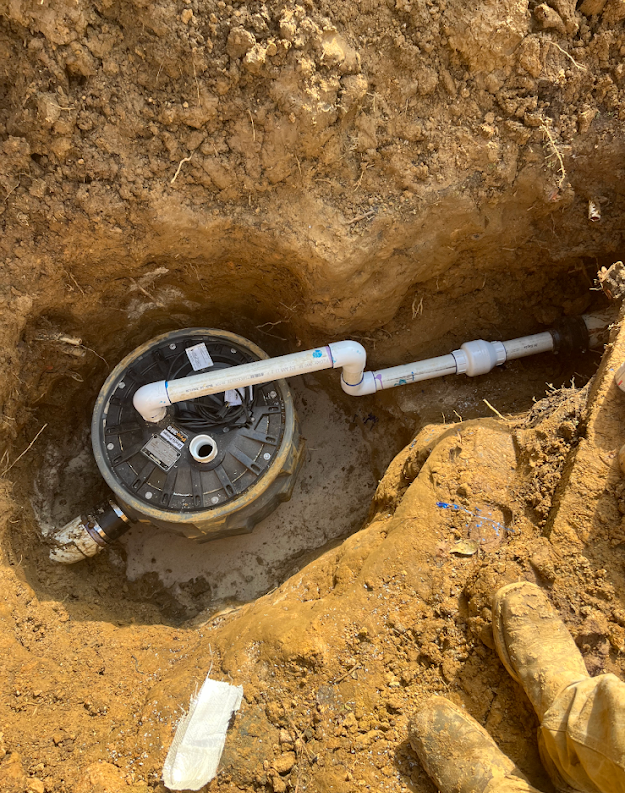 Open utility pit with a round meter box and white PVC piping in a dirt trench.
